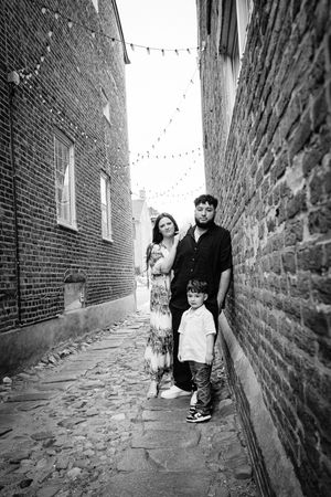 A family walks down a narrow cobblestone alley between old brick walls with string lights overhead in black and white.