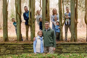 A group of people pose together against a stone wall and trees in an outdoor wooded setting.