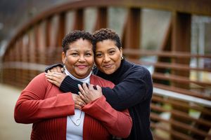 Two people embrace and smile warmly while standing on a wooden bridge outdoors.