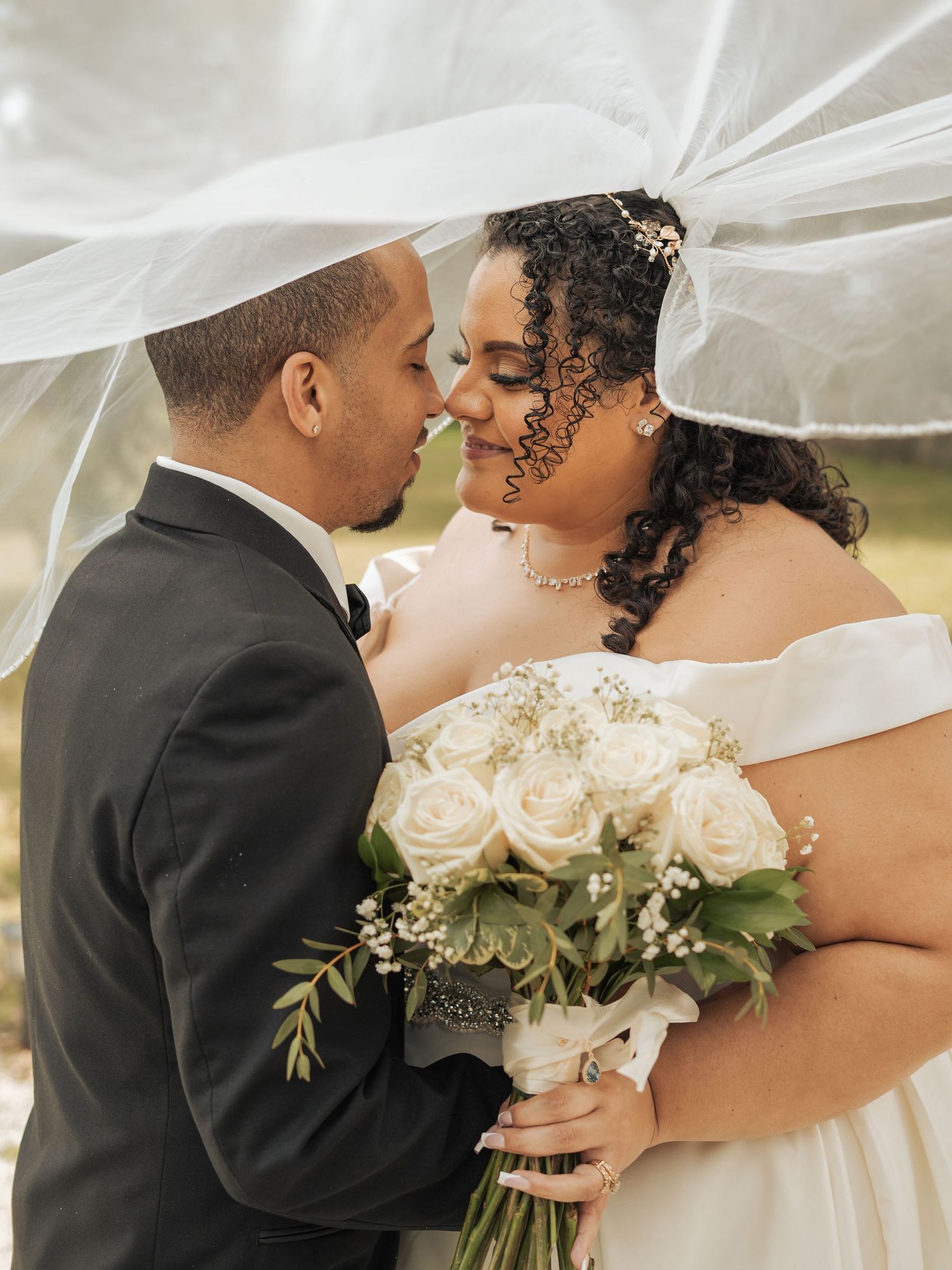 Romantic wedding portrait with a couple sharing an intimate moment under flowing white veil while holding white rose bouquet.