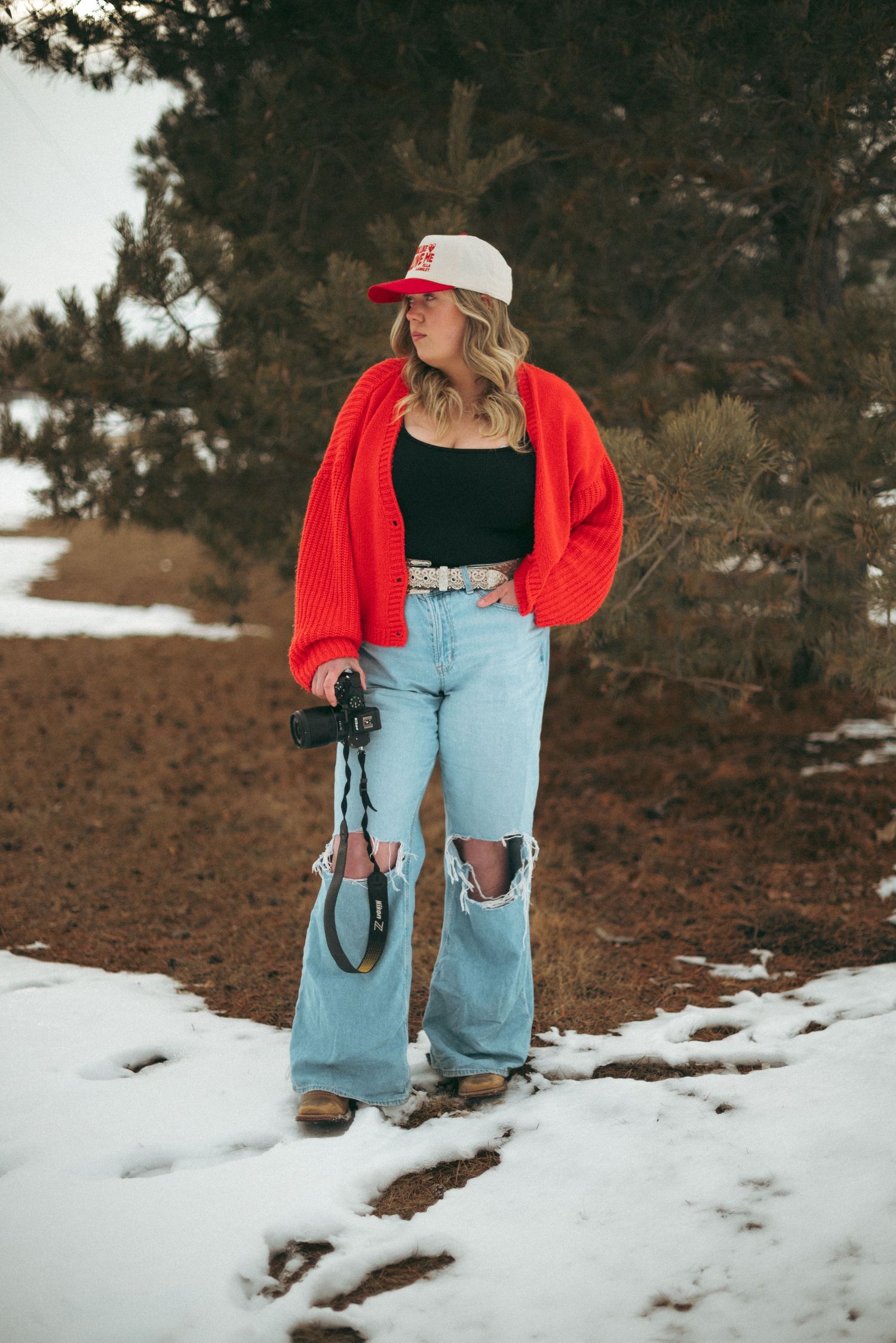 Person in red cardigan and ripped jeans poses outdoors in snowy winter setting wearing casual retro attire.