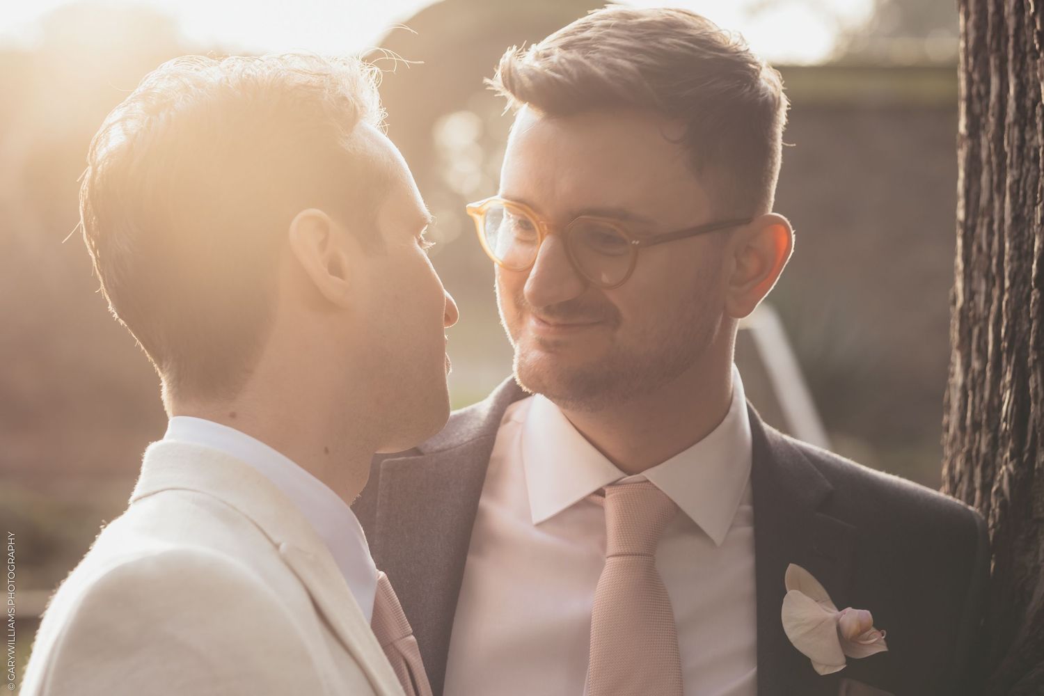 Two people in formal suits share an intimate moment at sunset with warm backlighting and a white boutonniere.