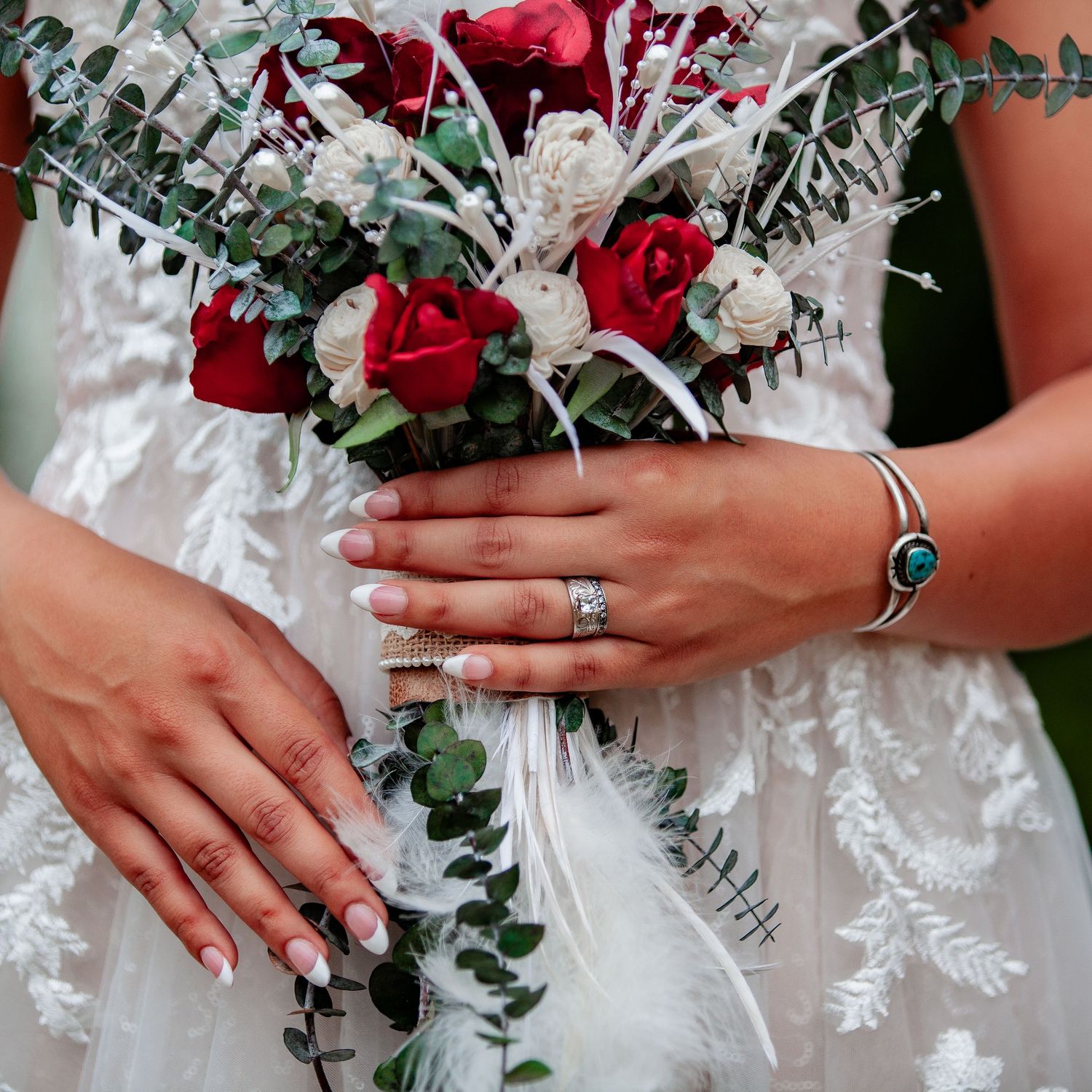 Close-up of hands holding red and white bridal bouquet with turquoise jewelry and wedding rings.