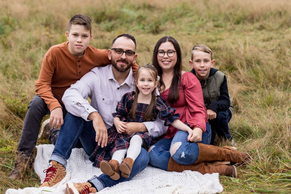 A family of five sits together on a blanket in a grassy field on a fall day.