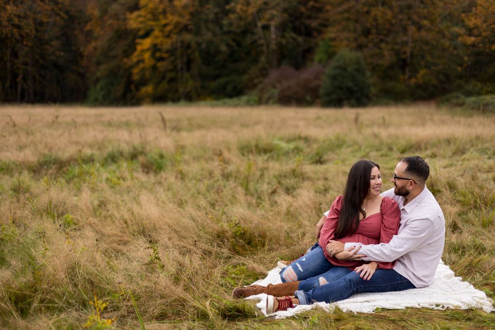 A couple sits on a blanket having a picnic in a golden autumn meadow near the forest edge.