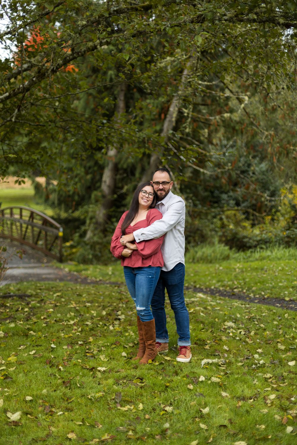 A couple embraces while standing on fallen autumn leaves in a park with trees and a wooden bridge in the background.