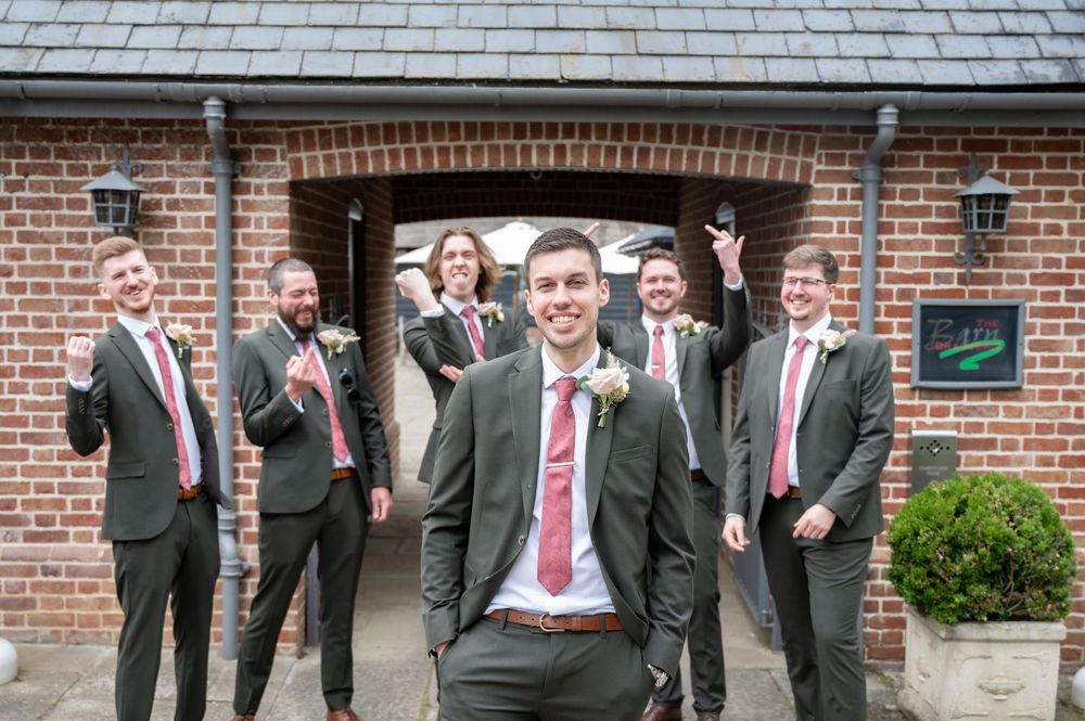 Group of groomsmen in suits pose cheerfully outside brick building entrance.