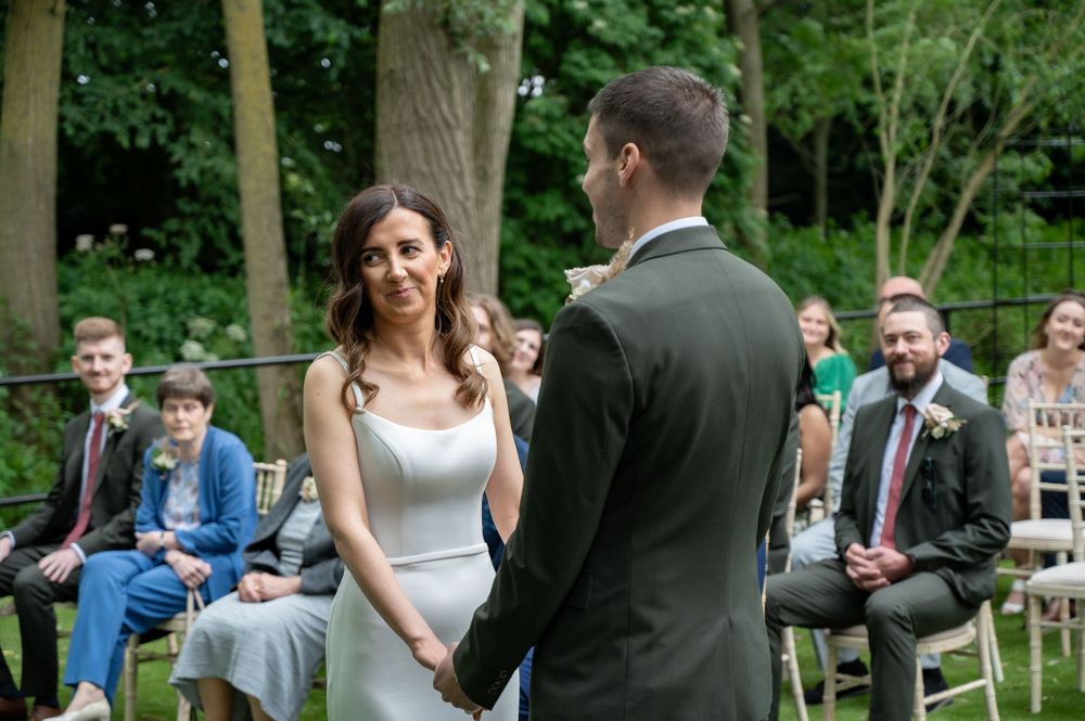Outdoor garden wedding ceremony with guests seated on chairs watching couple exchange vows.