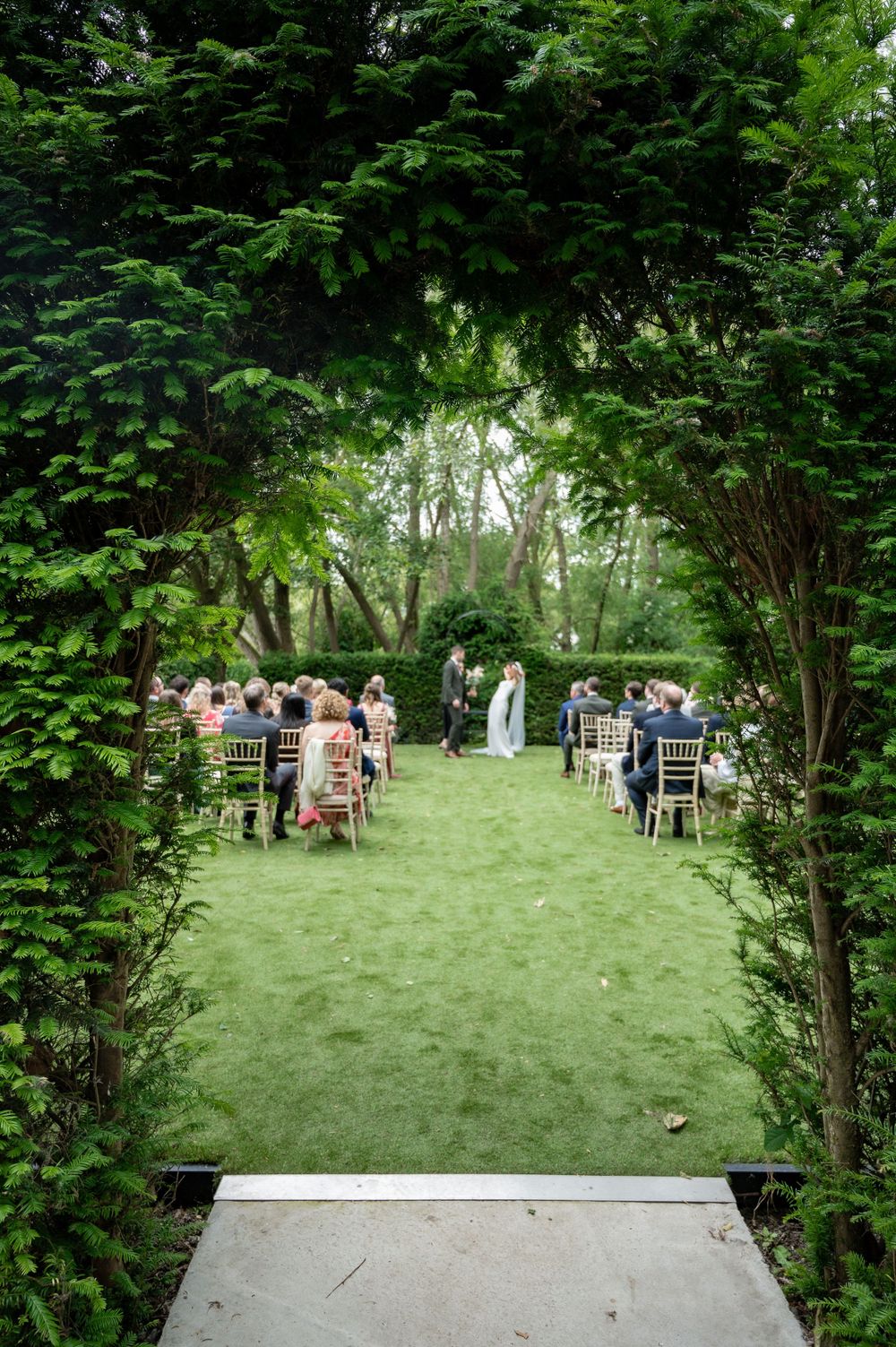 Garden wedding ceremony setup with white chairs arranged in rows beneath lush green archway.