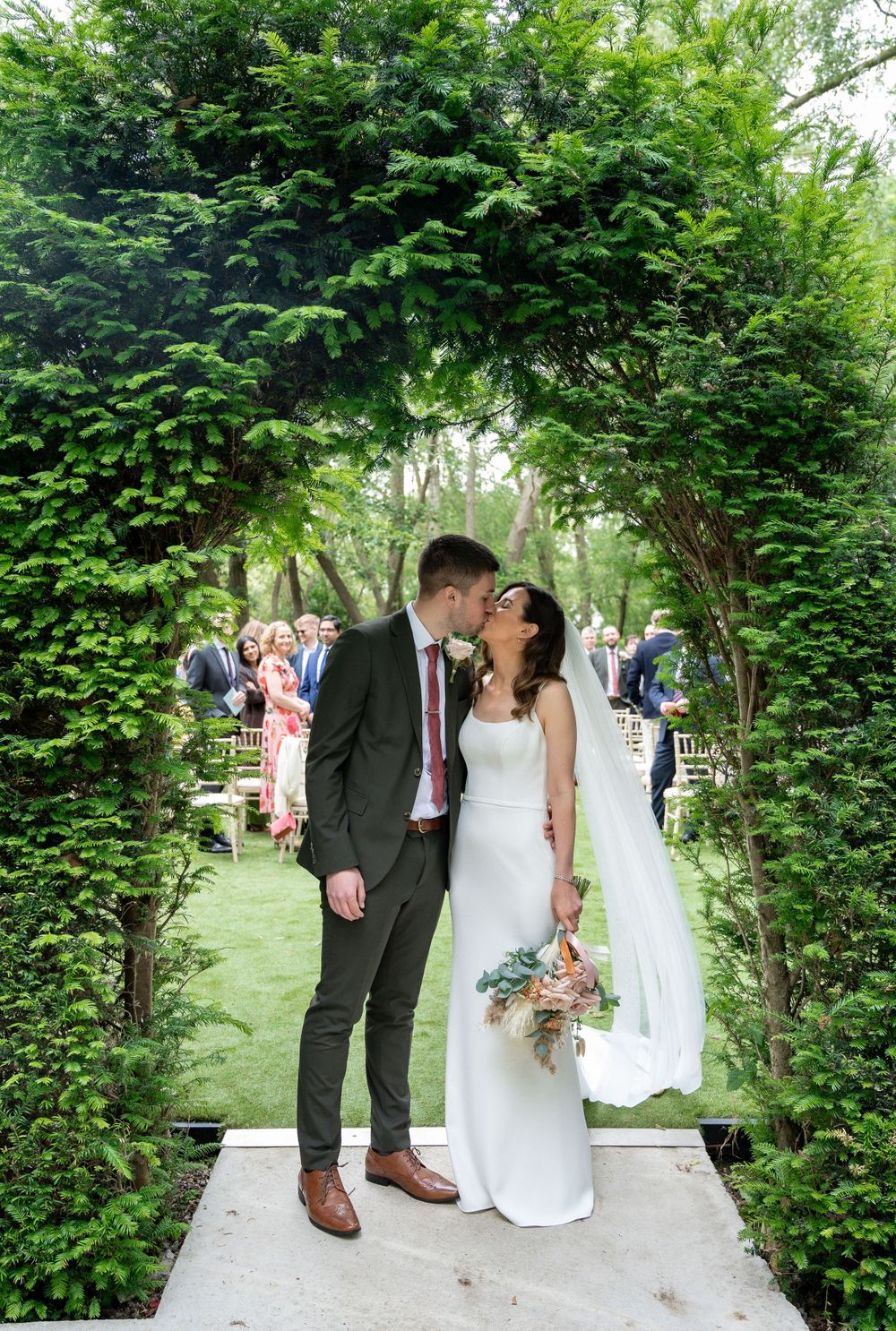 Newlyweds share an intimate moment under a lush green garden archway during an outdoor wedding ceremony.