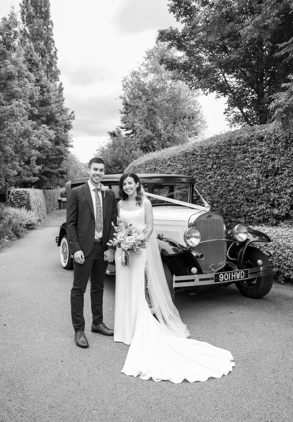 A wedding couple poses beside a vintage car in a scenic black and white portrait.