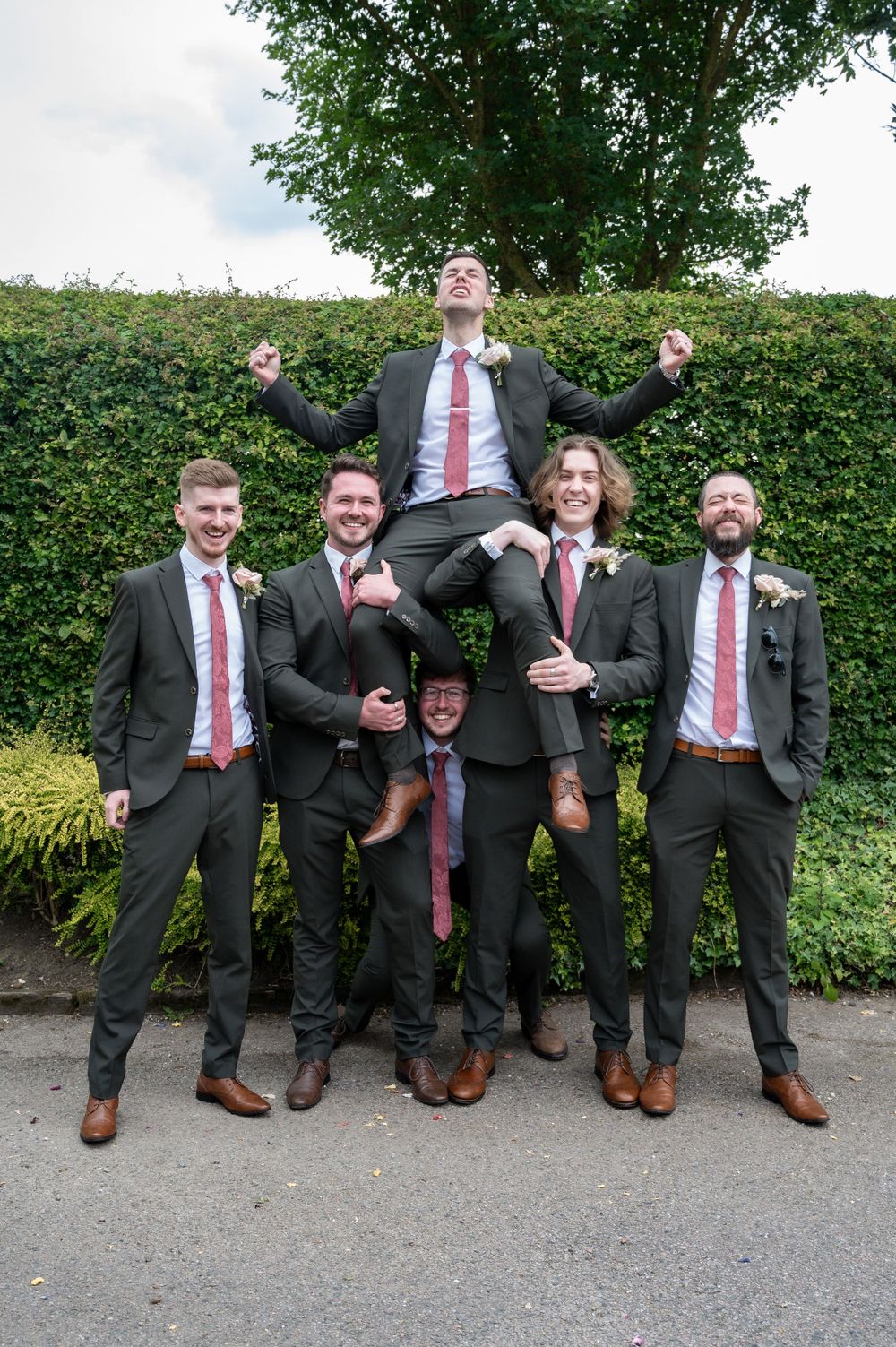 Groomsmen in matching suits and red ties perform a playful lift pose against a hedge backdrop.