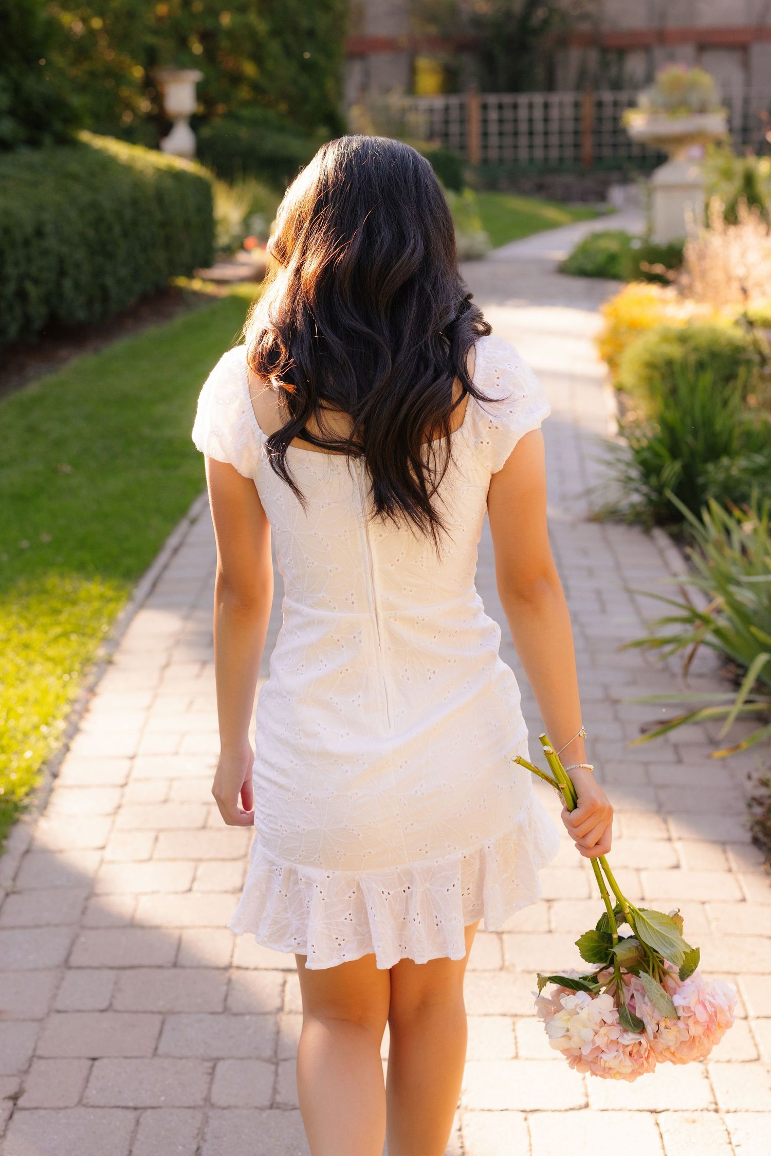 Back view of person in white summer dress walking along garden path holding flowers at sunset.