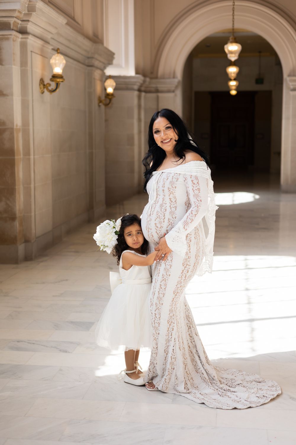 Pregnant woman in white lace gown poses with young flower girl in elegant marble hall.