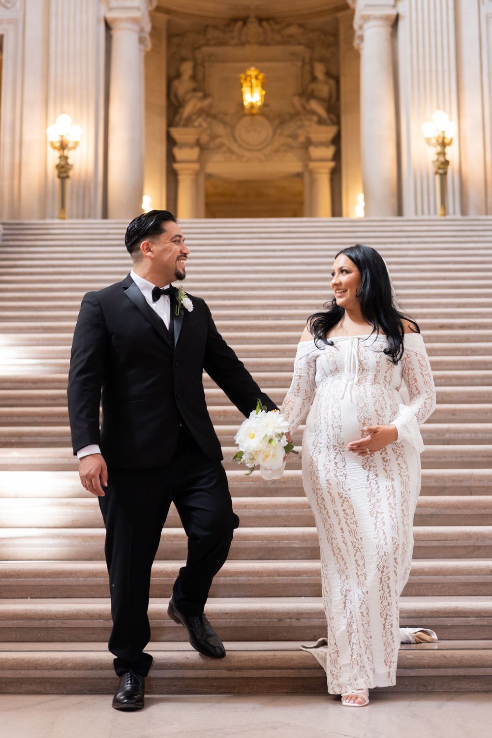 Pregnant bride in white lace gown and groom in black tuxedo walking down grand staircase holding hands.