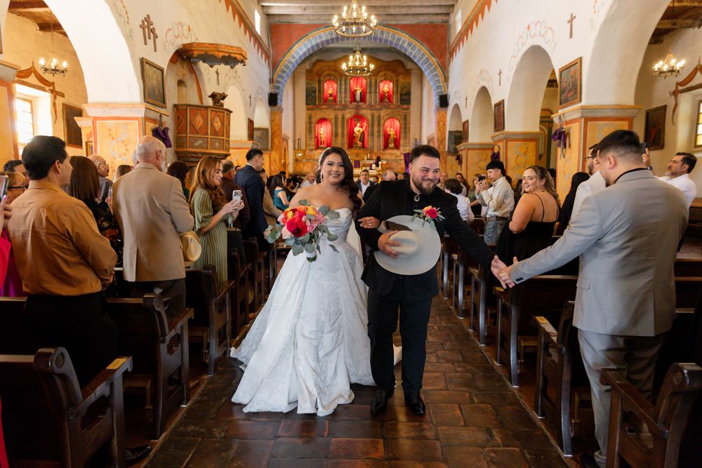 Bride and groom walk down church aisle after wedding ceremony, guests standing in wooden pews, colorful altar backdrop.