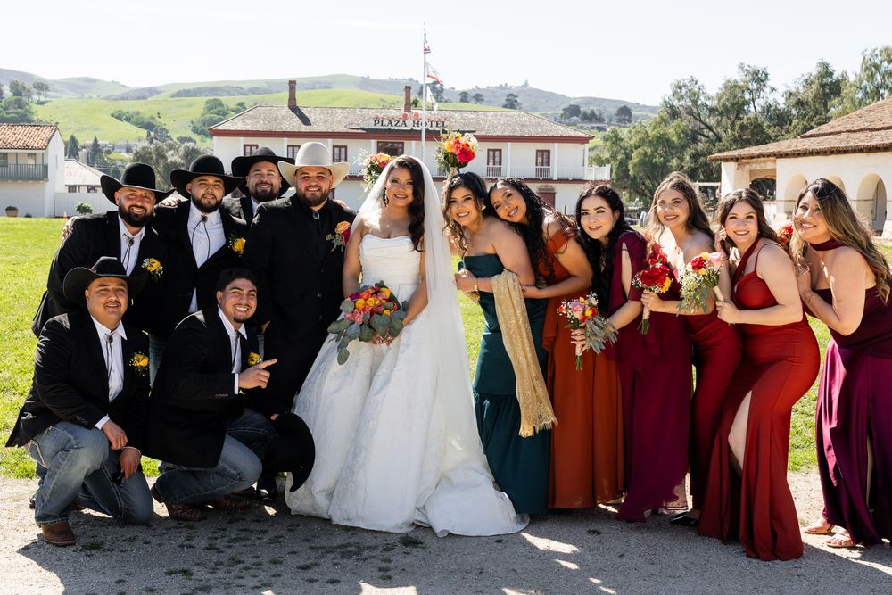 Wedding party poses outdoors with bride in white gown, groomsmen in black, and bridesmaids in red and green dresses.