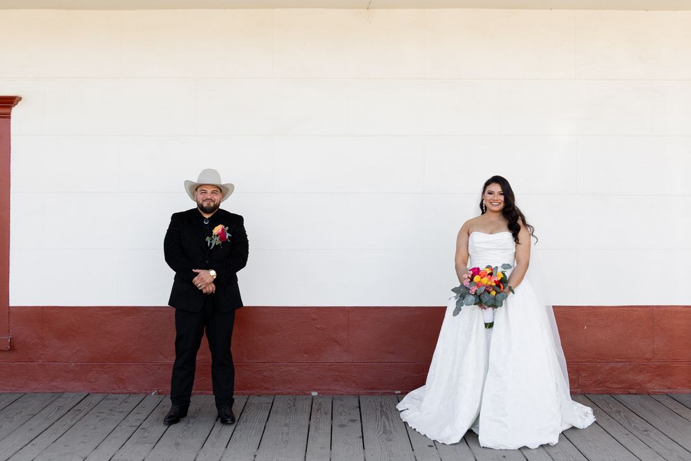 Bride in white gown holding colorful bouquet stands with groom in black suit and cowboy hat outdoors.