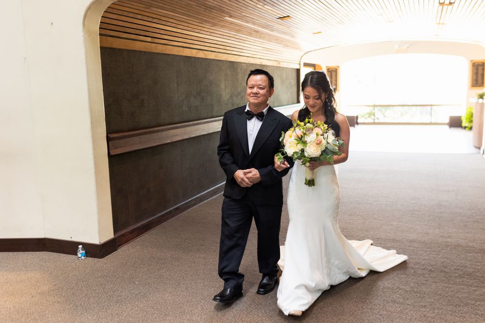 A couple walks down a carpeted hallway, with the bride carrying a bouquet of yellow and white flowers.