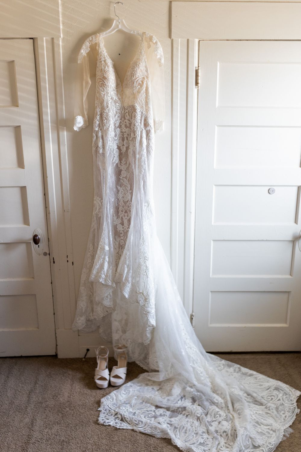 A delicate lace wedding gown with long sleeves hanging against white doors with shoes below.
