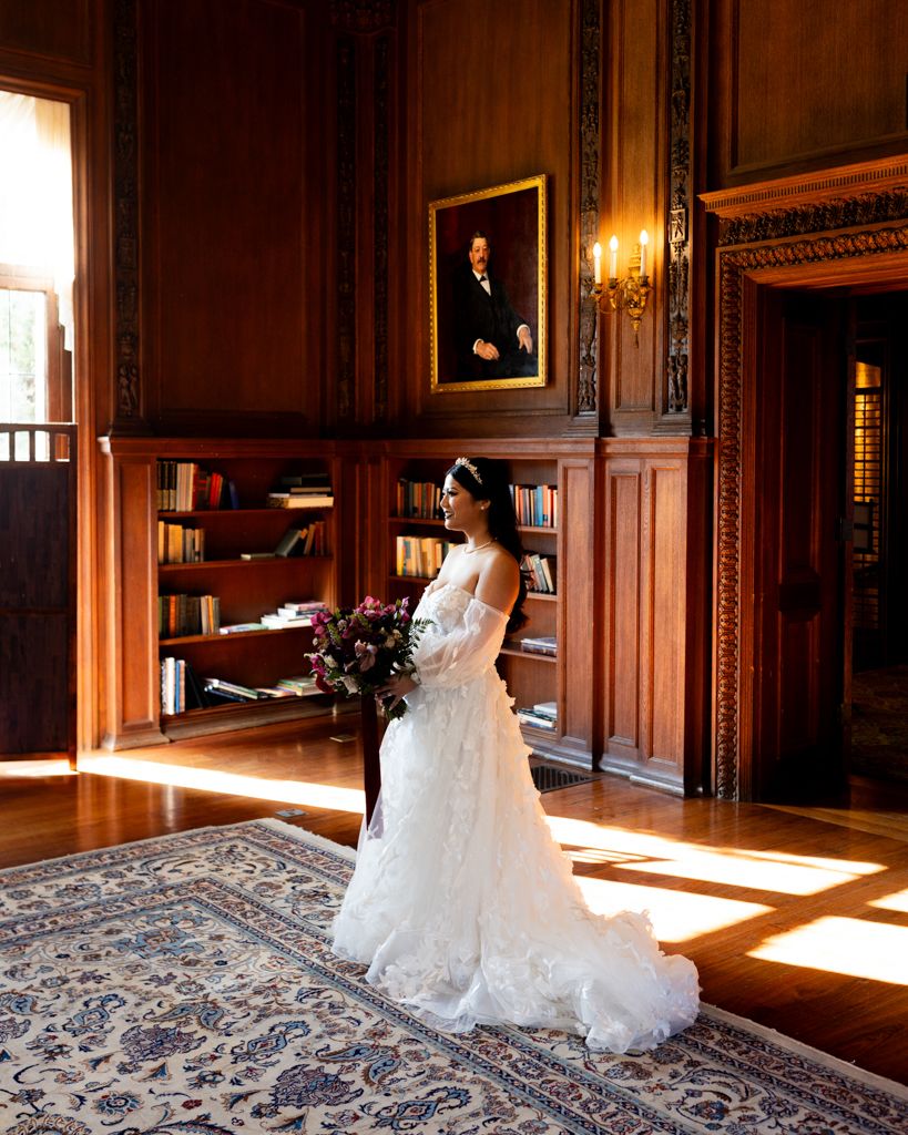 A bride in a lace gown stands in a wood-paneled library with dramatic window light.