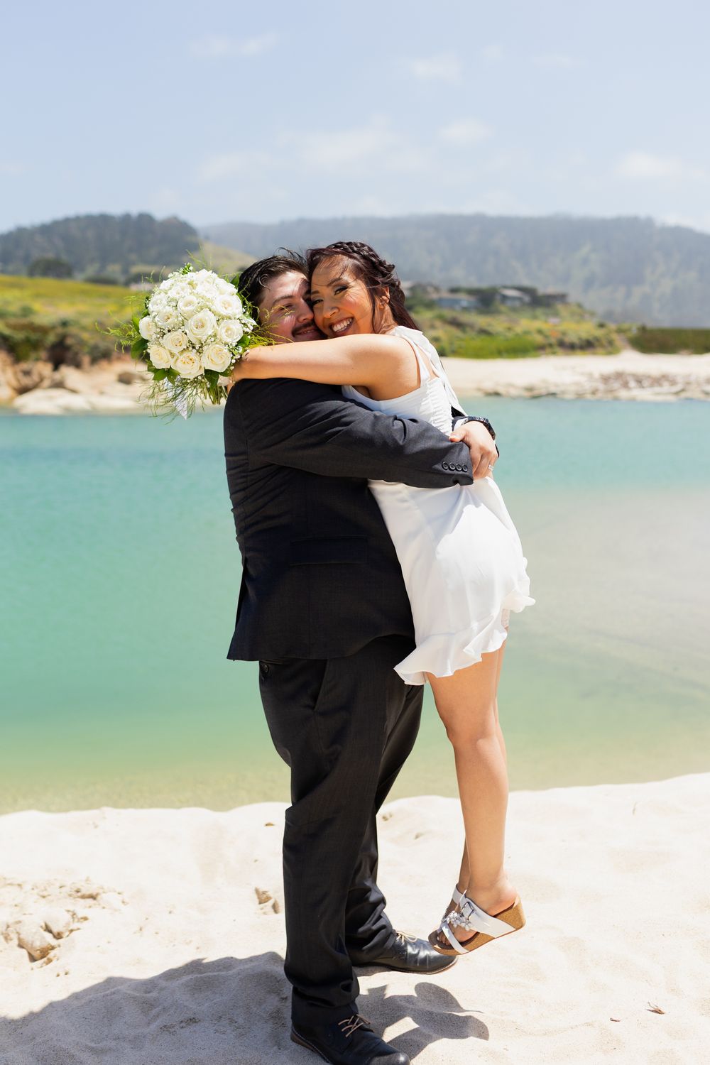 Newlywed couple embracing on sandy beach, bride in white dress holding bouquet, groom in black suit, turquoise water backdrop.