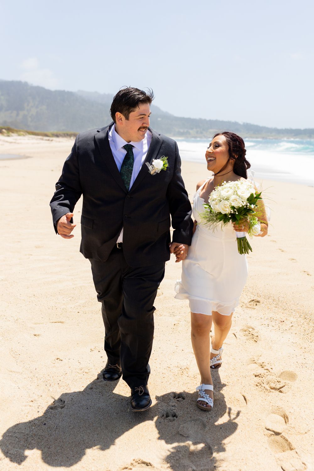 Happy newlyweds walking hand in hand on a sunny beach, bride in white dress holding bouquet, groom in black suit.
