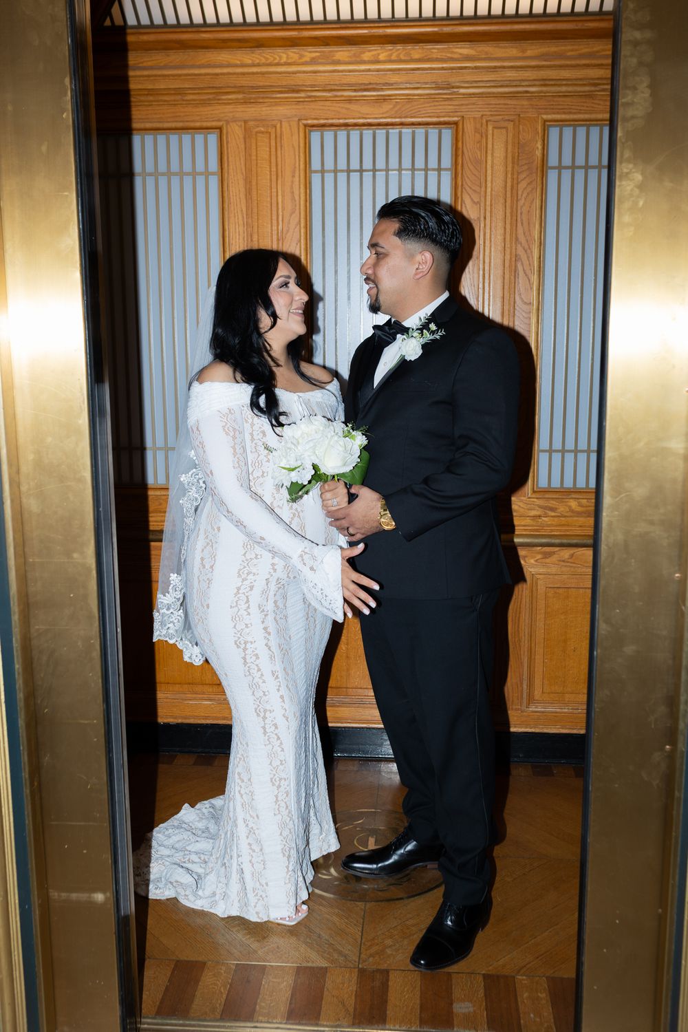 Bride in white lace gown and groom in black tuxedo share a romantic moment in an elegant elevator.
