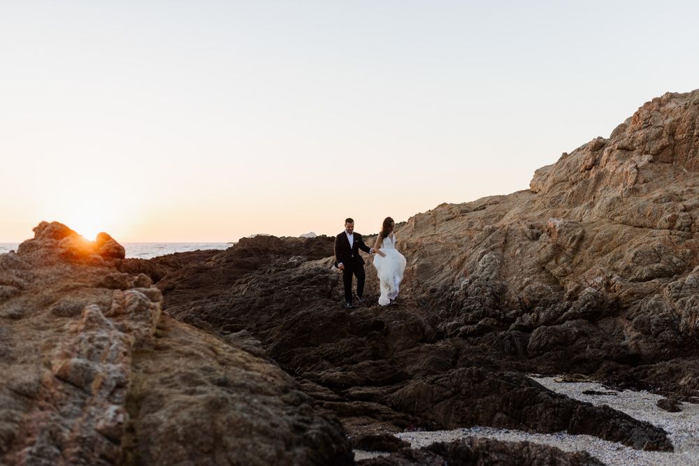 A couple silhouetted against a golden sunset on coastal rocks.