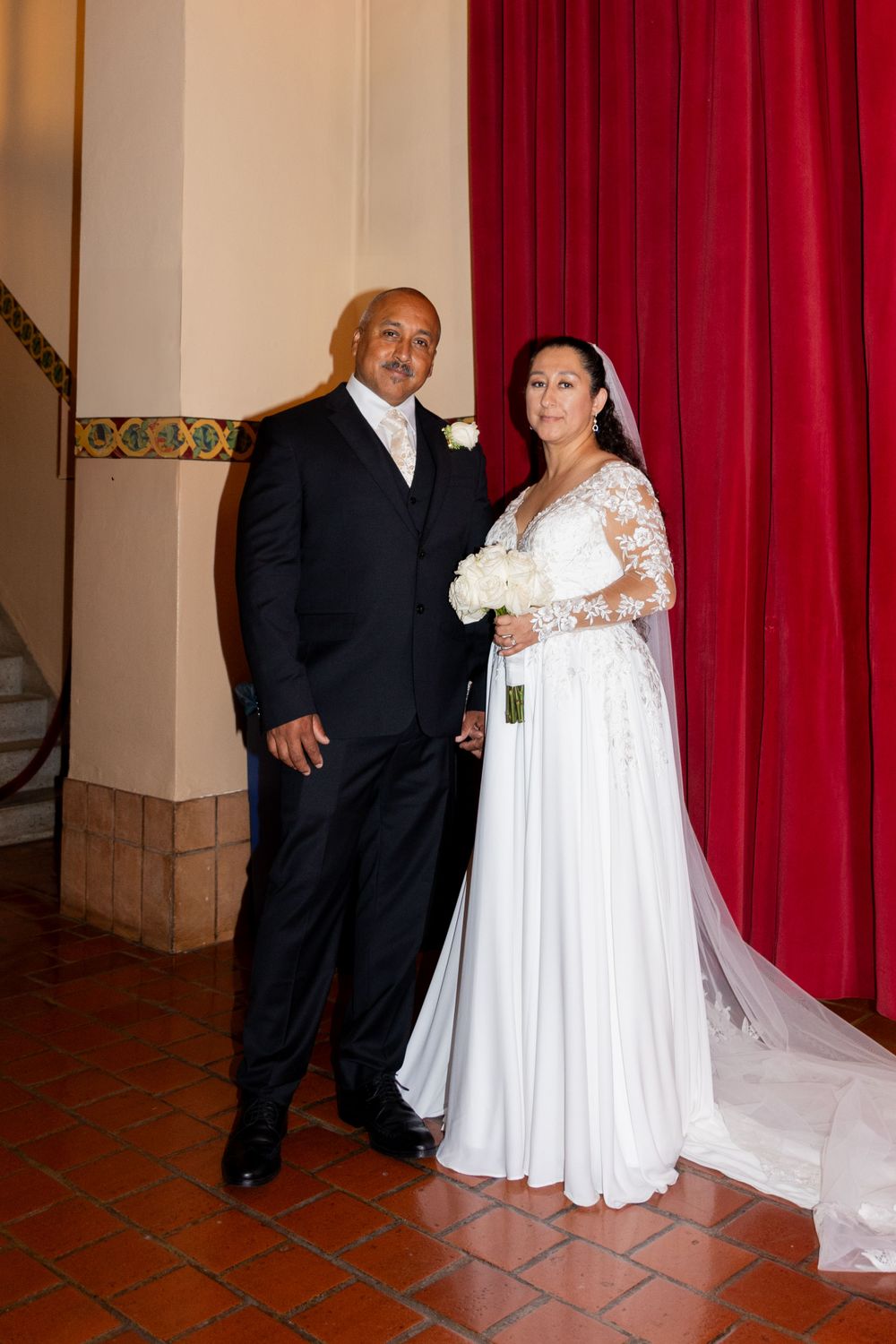 A couple in formal wedding attire poses together against a backdrop with red curtains and decorative wall details.
