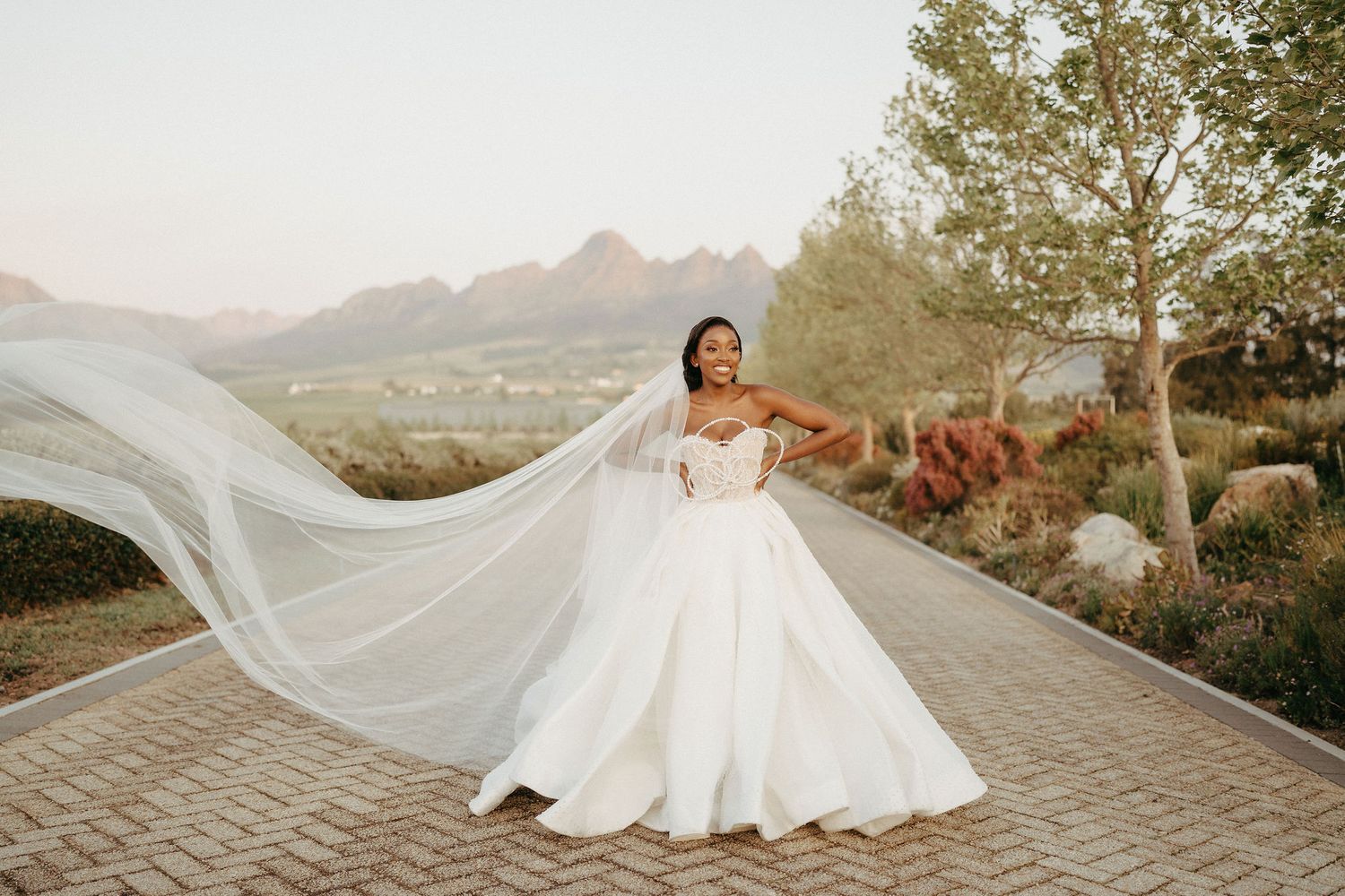 Flowing white wedding dress and veil billow dramatically against mountain landscape at sunset.