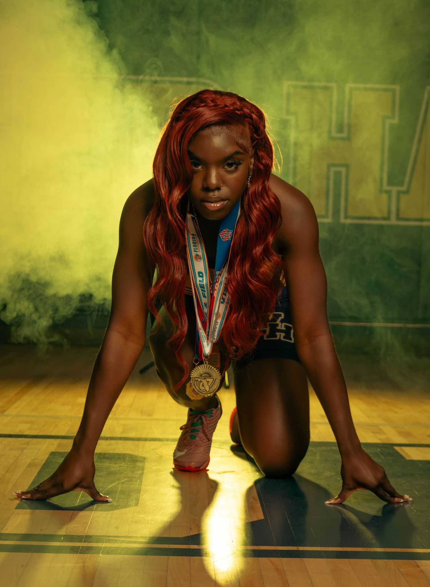 A female athlete in track uniform with gold medals poses in starting position on a gym floor.