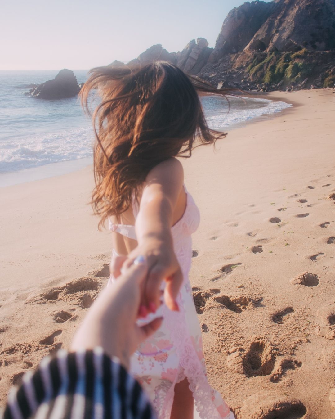 Brunette woman in pink floral dress leading someone by the hand along a sandy beach.