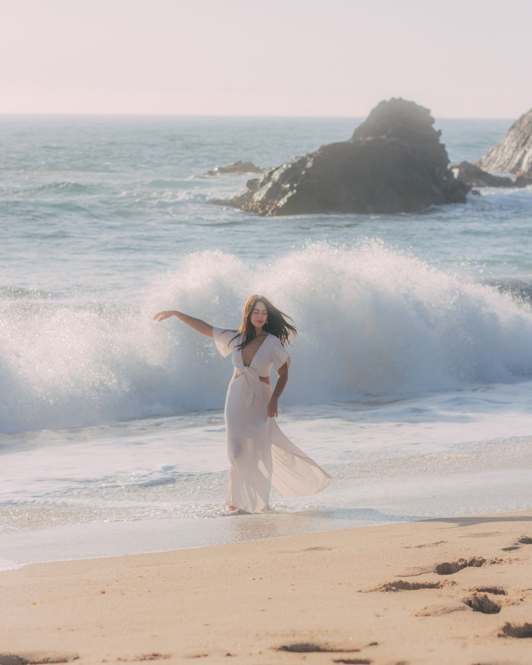 Woman in white dress dancing on beach with dramatic crashing waves and rocky coastline.