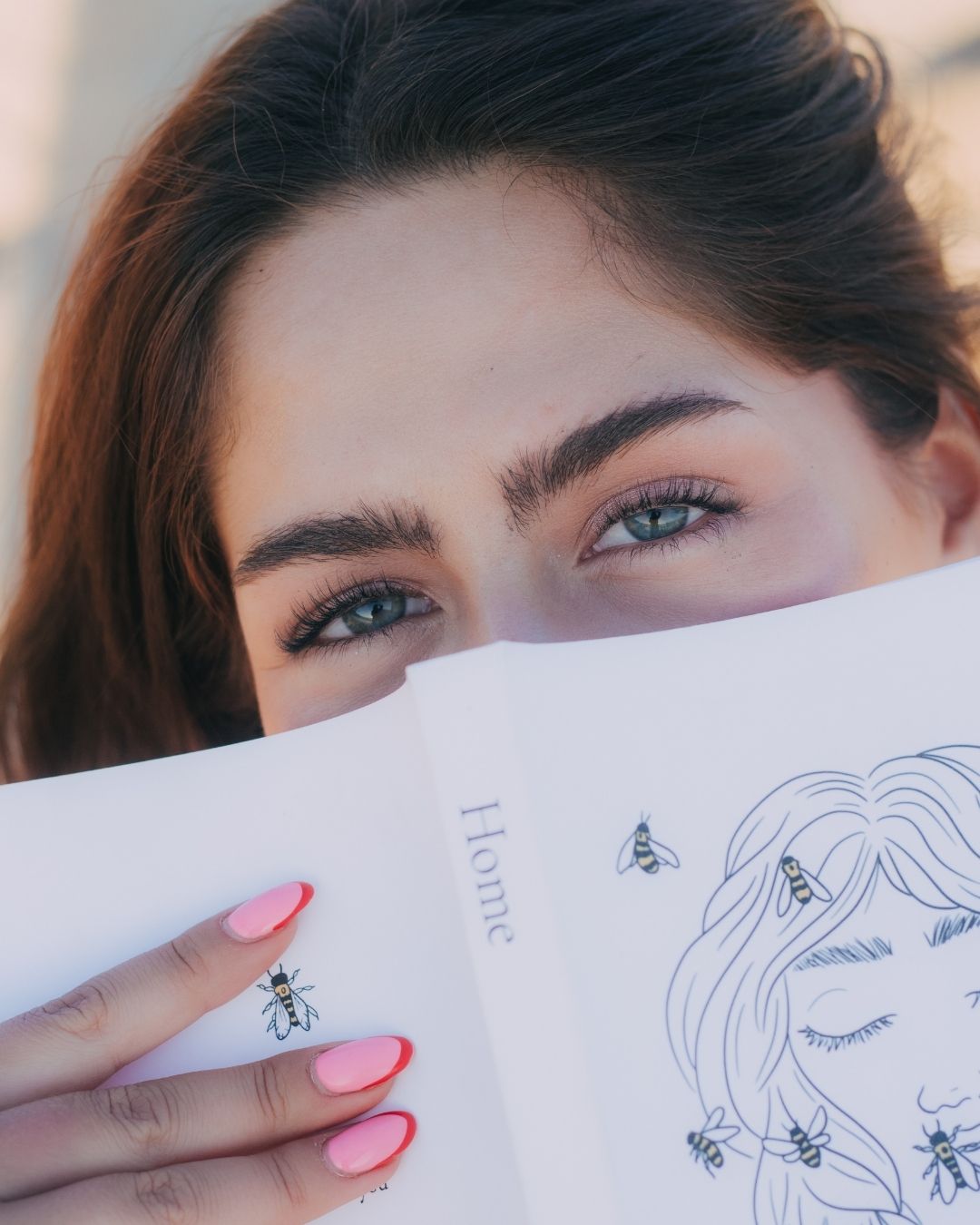 Close-up of woman with green eyes holding illustrated book with bee drawings and pink nails.