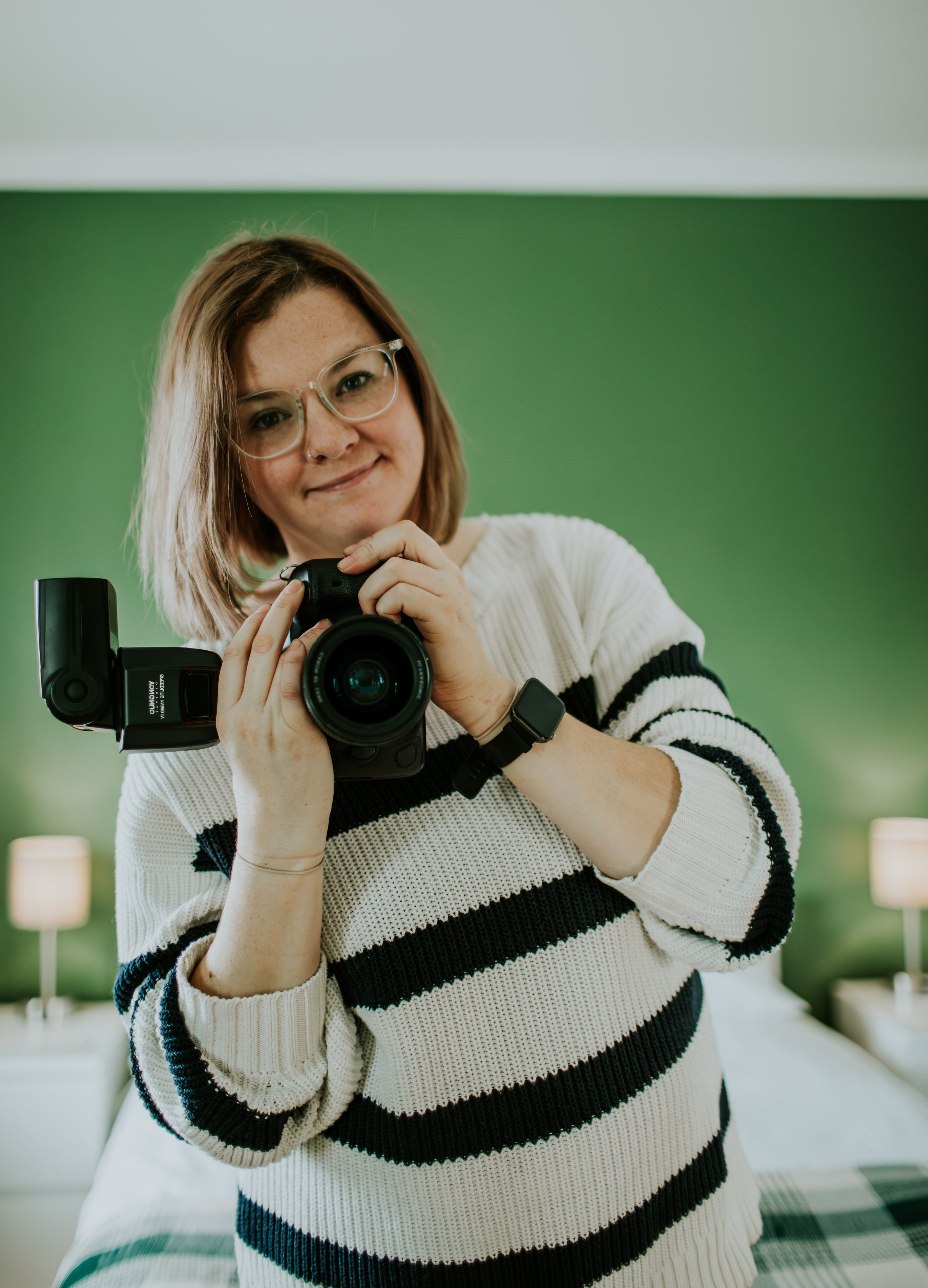 Photographer in striped sweater holds professional camera against green wall backdrop.