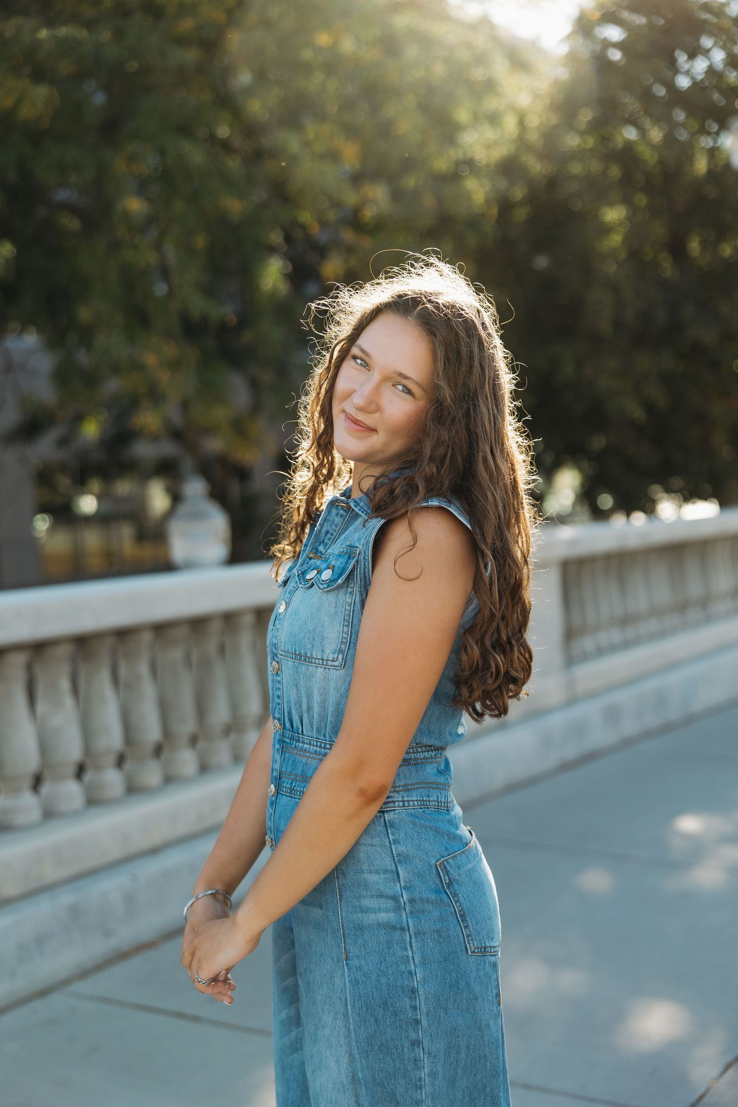 Person in denim outfit poses on pathway with sunlit trees and stone railing in background.