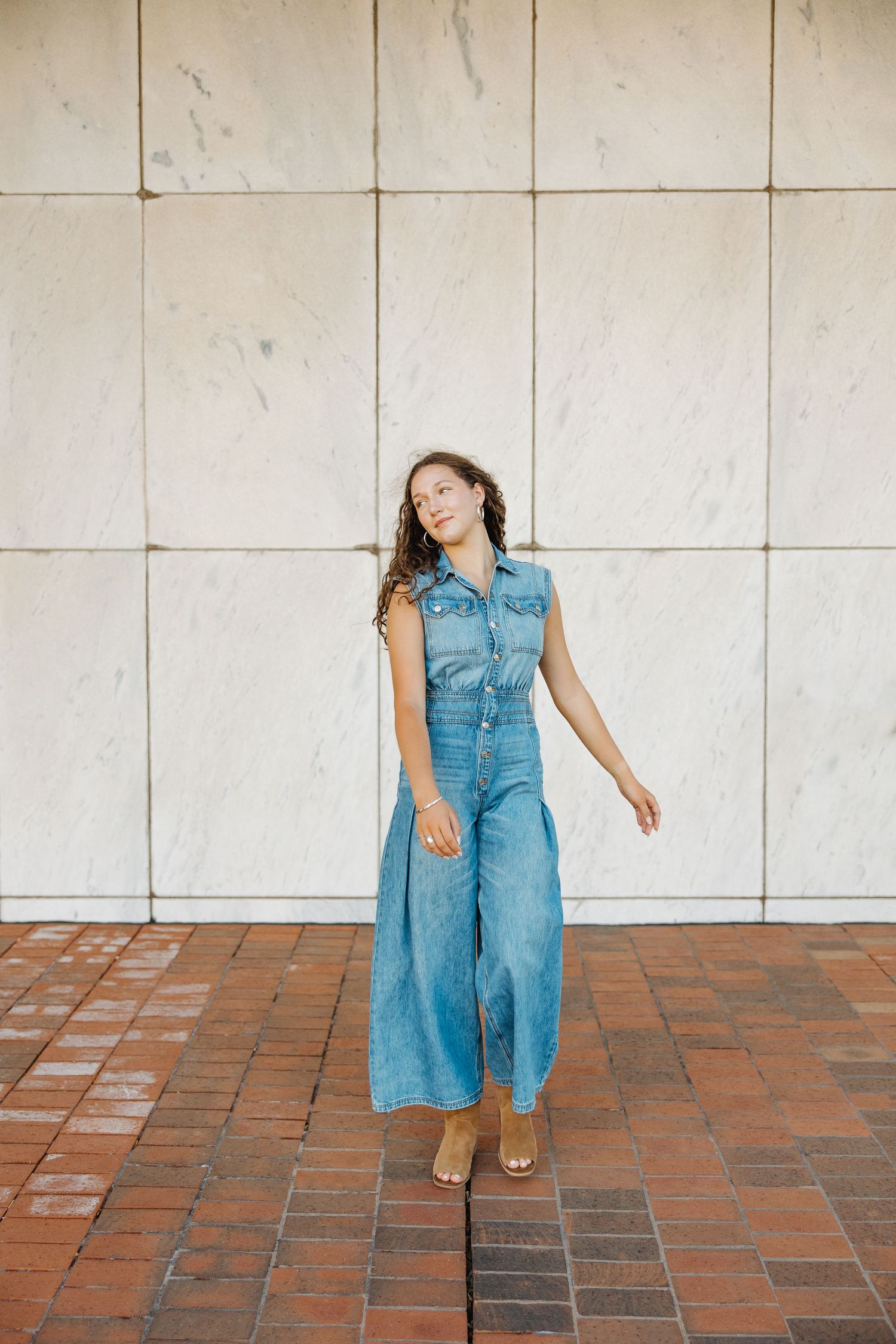 Person in blue denim jumpsuit poses against white tiled wall on brick pavement.