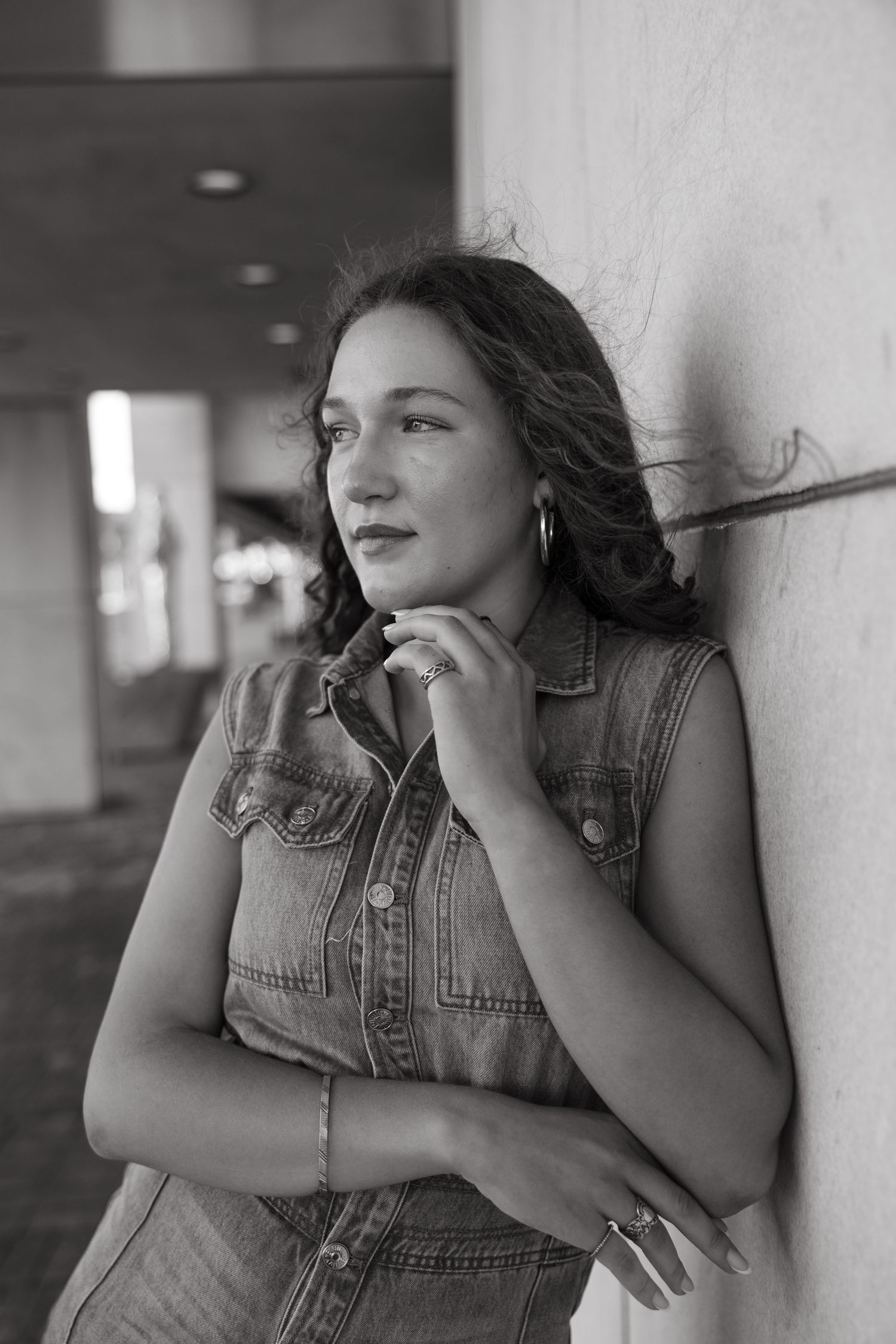 Black and white portrait of person in denim vest gazing thoughtfully against wall.