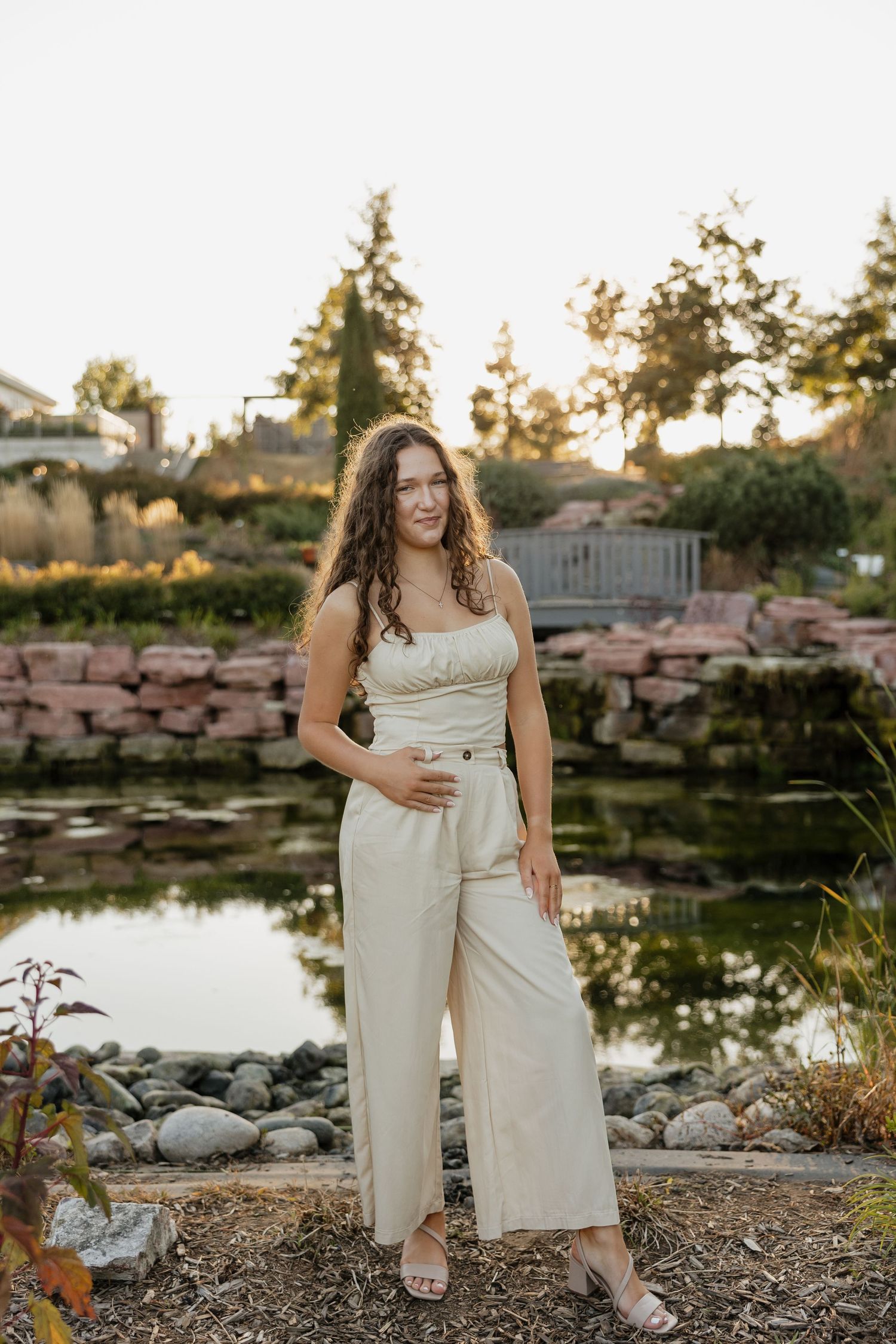 A person in white clothing poses by a decorative pond with stone landscaping at sunset.