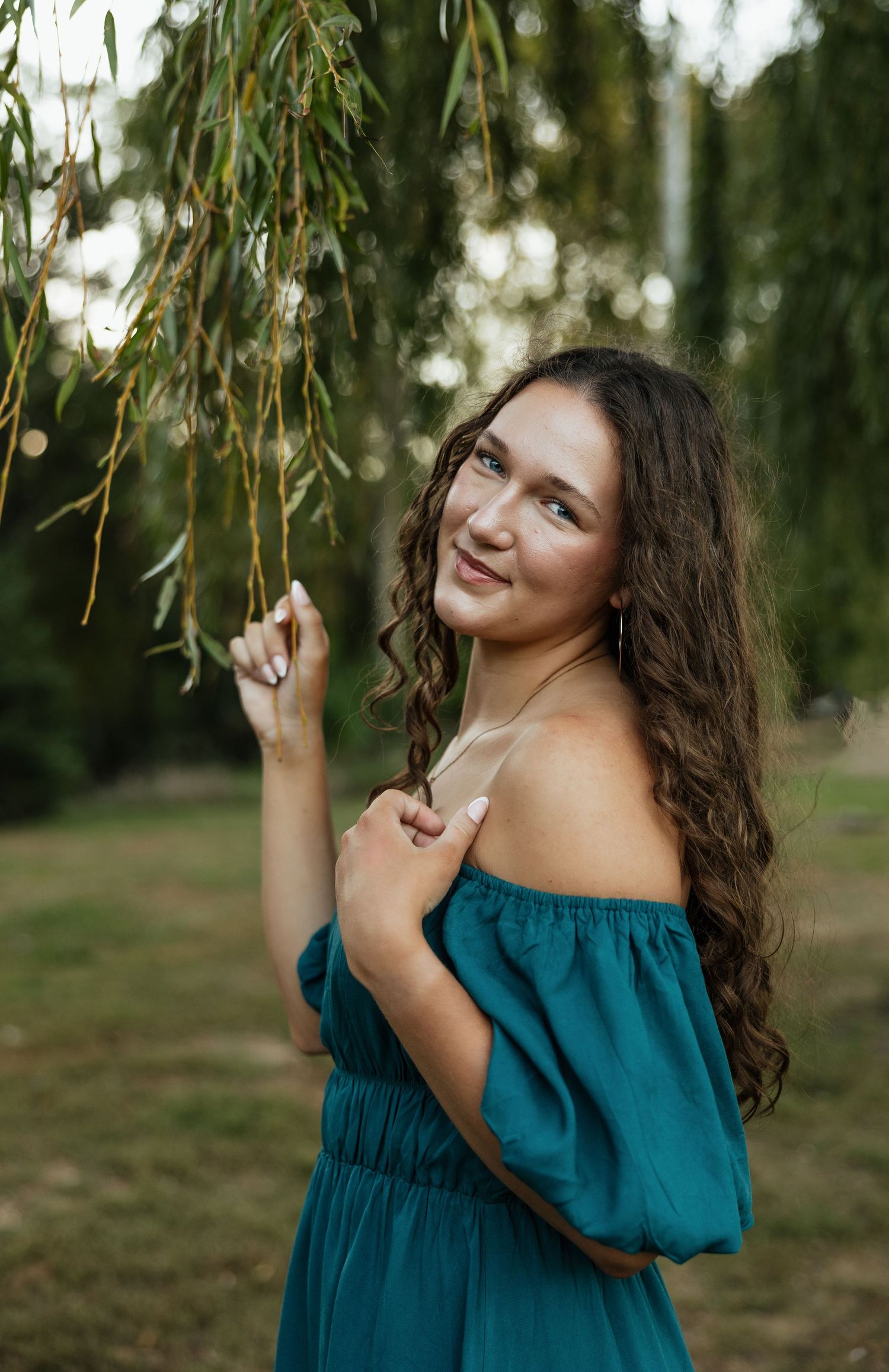 A portrait of someone in a teal off-shoulder dress standing beneath willow branches.