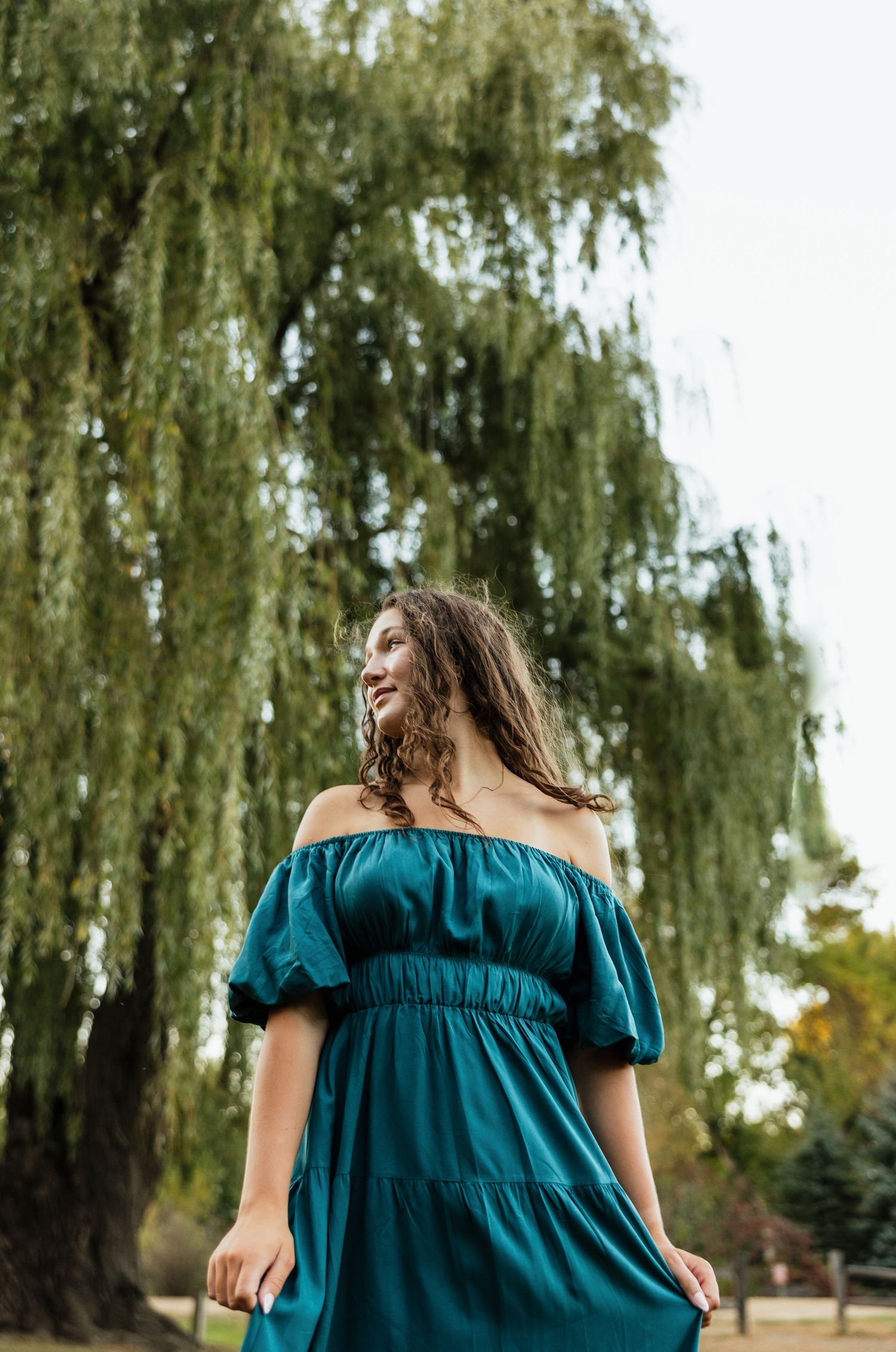 A low-angle shot of someone wearing a flowing teal dress under a weeping willow tree.