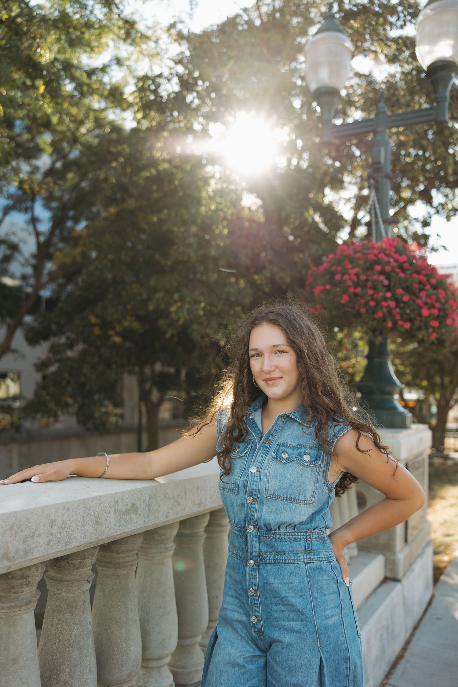Person in denim jumpsuit poses against stone railing with sunlit trees and lamp post in background.