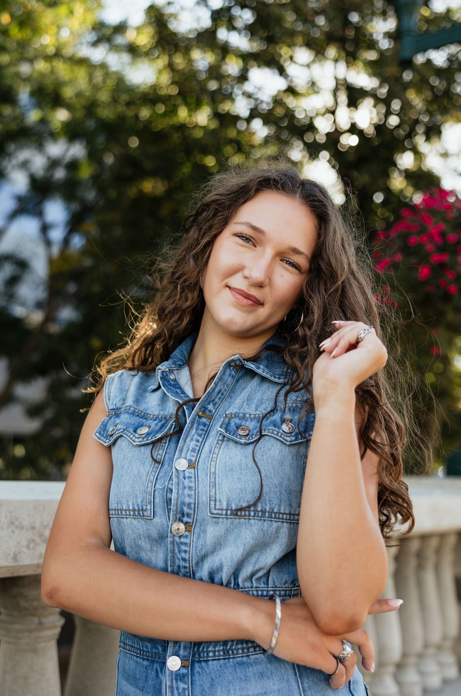Person in sleeveless denim top poses outdoors with natural backlit lighting and flowering plants.