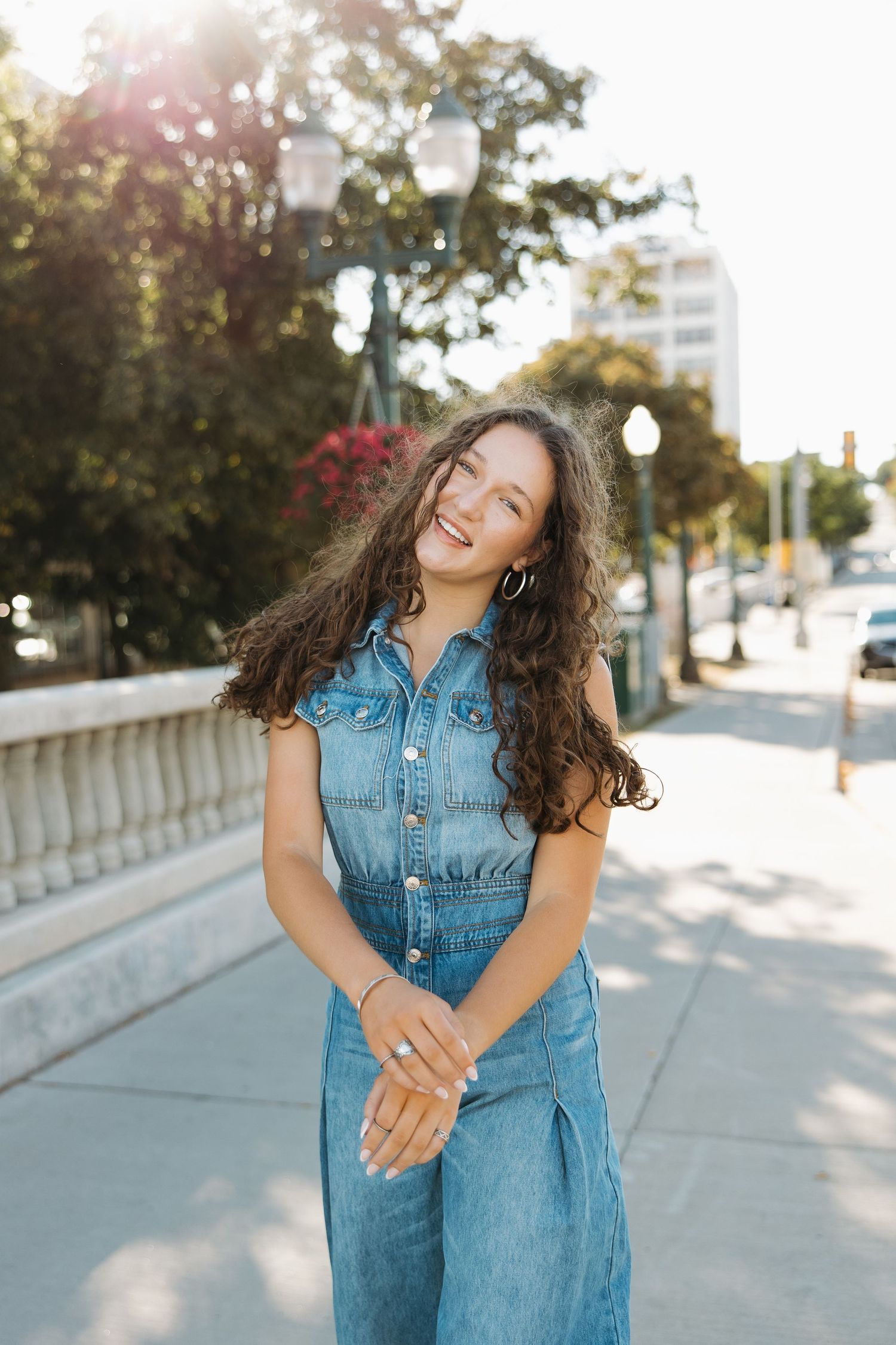 Person in denim jumpsuit smiling on urban sidewalk with street lamps and buildings in background.