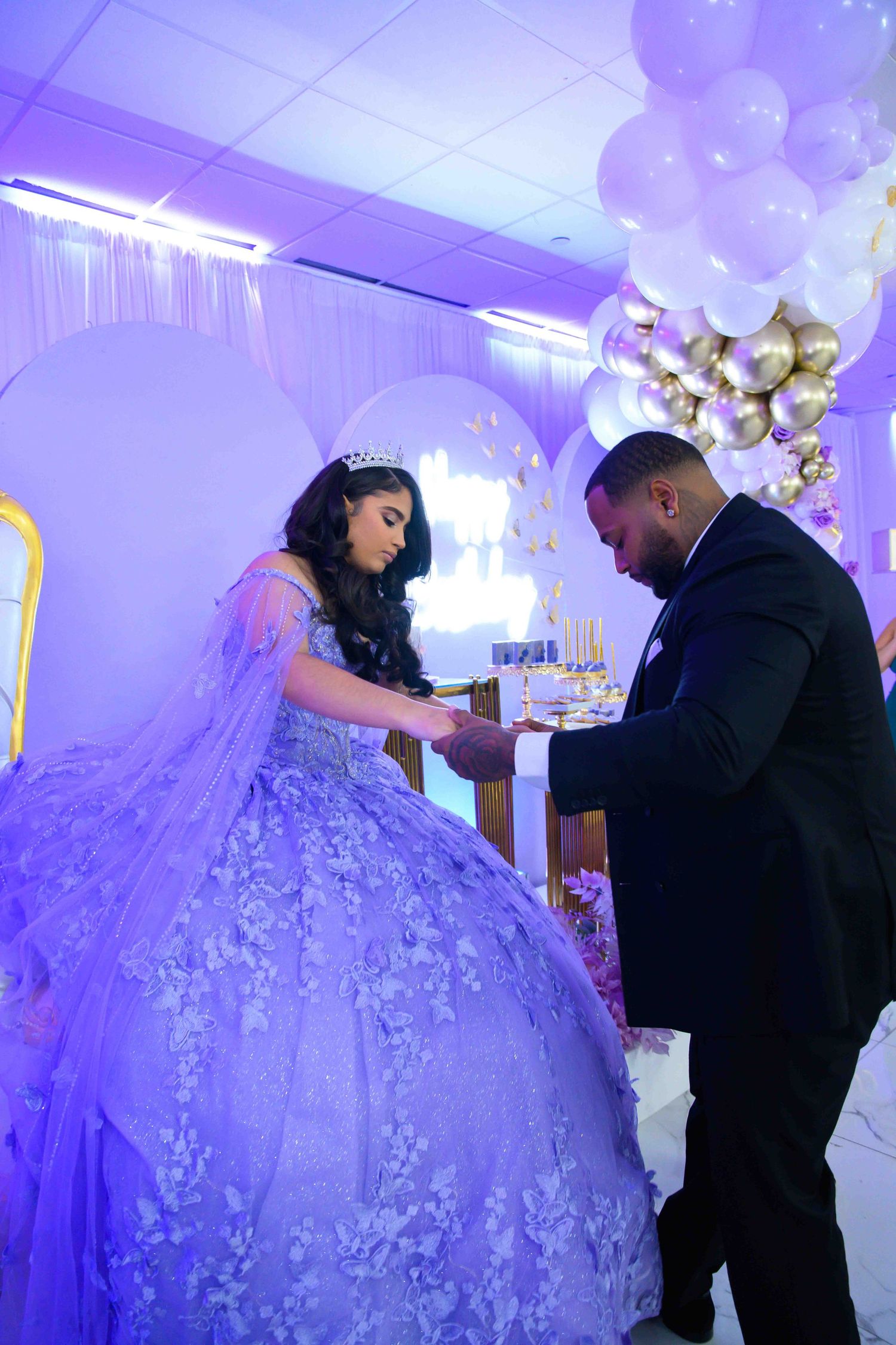 A couple in formal attire shares a moment under purple lighting with balloons and decorative elements.
