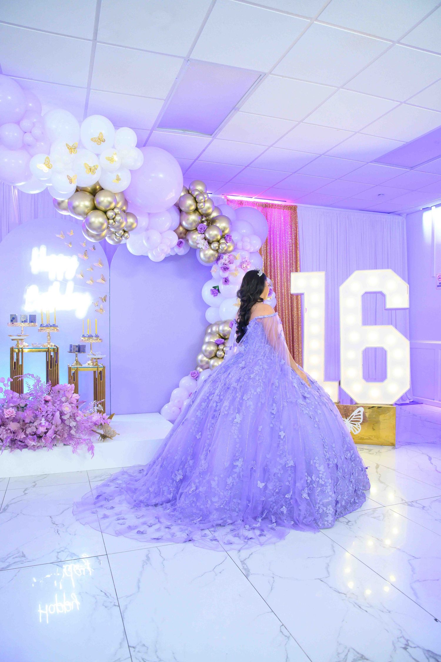 A lavender quinceanera ballgown in an elegant purple-lit room with white balloon arch and large illuminated number 16.