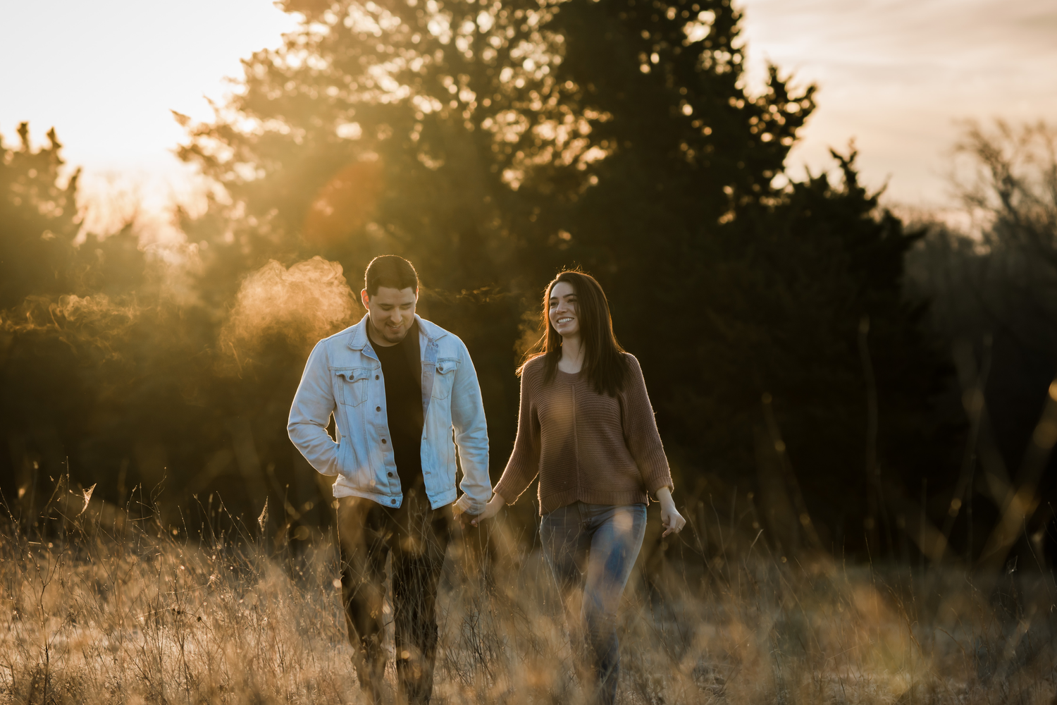 A couple walks through a field during golden hour with sunlit trees in the background.