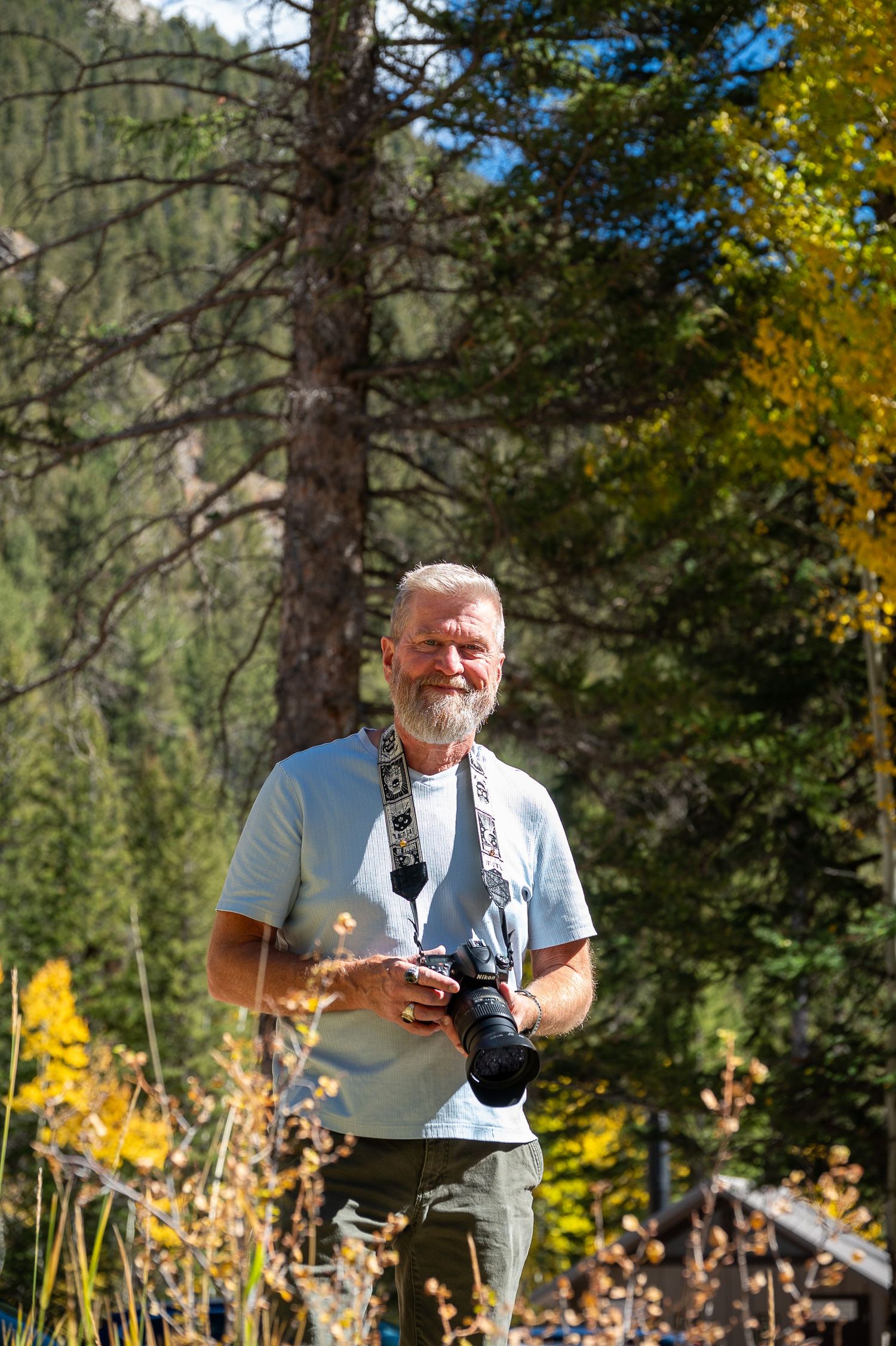 Photographer holding camera outdoors with autumn foliage and pine trees in background.