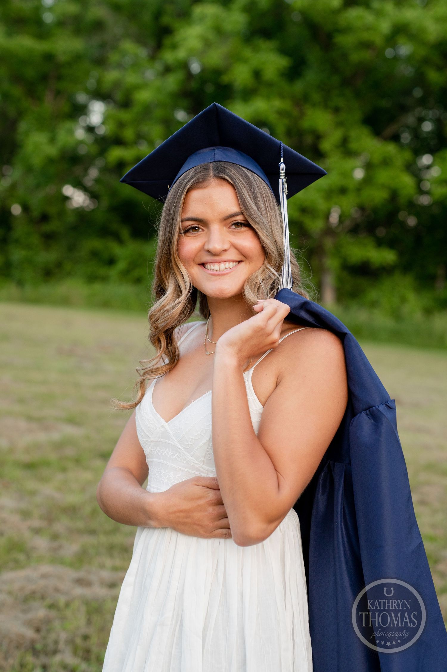 Graduate in white dress and navy cap holds graduation gown while posing outdoors.
